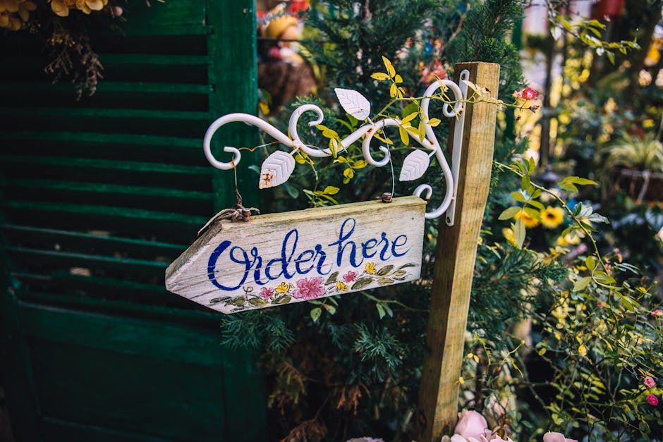 A close-up of a decorative garden sign hanging from a white wrought iron hook, with the word 'Order here' painted in blue cursive on a weathered wooden plank, decorated with pink and yellow floral mot