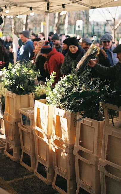 A busy outdoor flower stall under a canopy with several beige plastic crates filled with lush green floral arrangements, featuring a variety of foliage and delicate white flowers. Customers, dressed w