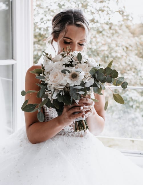 A bride holding a large floral bouquet featuring white roses, white daisies, and eucalyptus leaves. The arrangement is wrapped with a white ribbon, and she is wearing a white lace wedding dress. She i