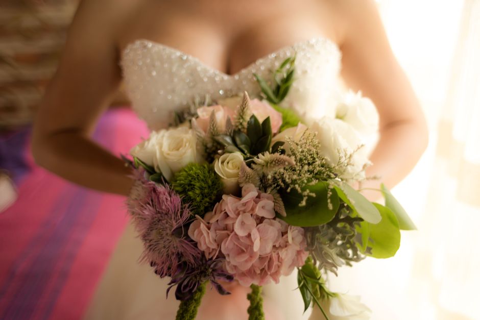 A bride holding a bridal bouquet featuring a mixed arrangement of fresh flowers, including soft pink hydrangeas, white roses, and green foliage, with some purple and white accent blooms. The bouquet i