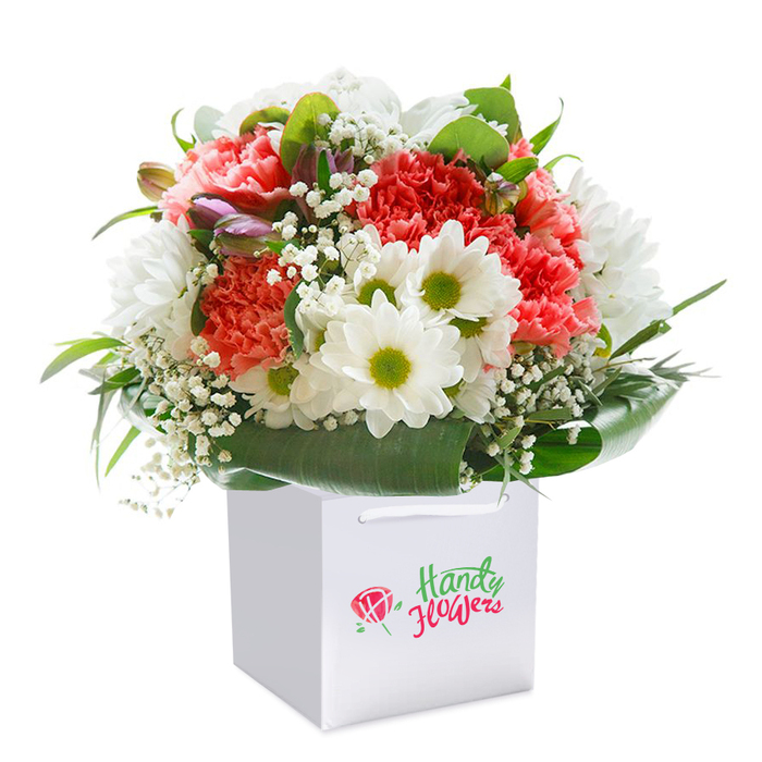 Presented in a crisp, square white gift bag with strong ribbon handles, this vibrant bouquet captures the gentle elegance of a Surrey Quays morning. Artfully arranged by local florists, the central blooms-lush coral and peach-hued carnations-unfurl their abundant, ruffled petals, evoking a sense of movement and inviting touch. Alongside them, fresh white daisies with luminous yellow-green centers impart a playful brightness, echoing the cheerful mood of the bustling markets near Canada Water. Clouds of white baby's breath introduce a whisper-light delicacy, weaving between the larger flowers and emphasizing the arrangement's airy sophistication. Adding further dimension, a few closed buds tinged with purple and pink, perhaps early spring alstroemeria, nestle discreetly on the bouquet's side, hinting at petal layers yet to reveal themselves. All these elements are gathered within a circle of bold, glossy green leaves, whose broad surfaces give structure and frame the vibrant palette above. The overall shape is rounded and full, suggesting plenty and care, while the bouquet stands proud against a pure white background, highlighting each flower's freshness and detailed handwork. The subtle logo printed on the front of the bag quietly reflects local expertise and service. Ideal for gifting-whether for a milestone, spontaneous gesture, or Mother's Day surprise-this bouquet resonates with the easy sophistication and neighbourly warmth that define life in Surrey Quays. Its artistry and thoughtful design embody the tradition of impeccable, heartfelt floristry found in this riverside community.