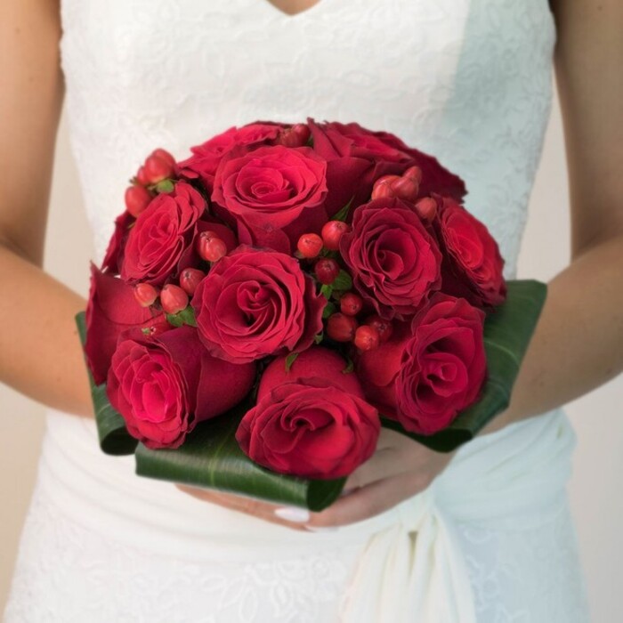 This evocative image, set in a tranquil corner of Surrey Quays, captures a close-up, waist-up view of a bride holding a rounded bouquet brimming with deep, velvet red roses. Meticulously arranged so each flower gently presses against its neighbours, the bouquet draws the eye with its rich gradients of red-some petals forming tight, secretive whorls, others gently peeling open to suggest hope and devotion. Clusters of glossy red hypericum berries peek out among the roses, their polished skins a playful and elegant accent that invites both sight and touch. At the bouquet's base, broad emerald green leaves wrap gracefully, their folded edges echoing the careful lines of a florist's practiced hand-much like arrangements you might find near Surrey Quays Road or Market Square. The bride's hands, manicured and delicate, anchor the composition, while a sliver of her flowing, lace-detailed white dress drifts into view from the lower edge, bathed in a soft, natural glow. The subtle, pale background ensures full focus remains on the floral artistry, evoking feelings of romance and gentle celebration ideal for Surrey Quays weddings. Created by Flowers Surrey Quays, this bouquet celebrates the area's spirit-traditional yet thoroughly contemporary in its beauty.