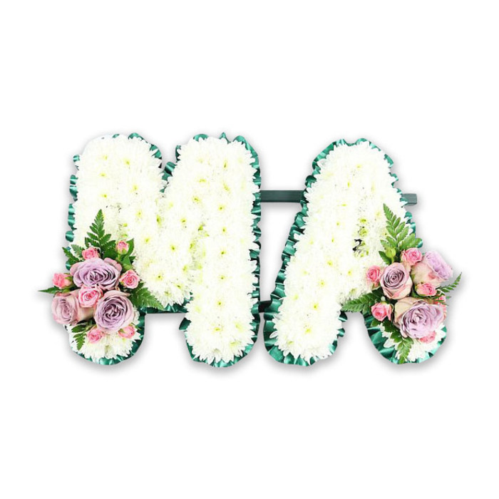 The photograph presents a refined funeral floral tribute that spells the letters MA, composed with the kind of precision and tenderness often requested for services around Surrey Quays and the nearby Rotherhithe churches. Both letters are formed from a sturdy backing that disappears beneath a dense layer of white chrysanthemums, each bloom pressed closely to its neighbour to create a plush, seamless surface. The chrysanthemum petals are small and slightly curved, overlapping to give a slight ripple effect, while the overall visual impression remains one of calm, steady whiteness-symbolic of honour, innocence, and peace. Encircling each letter is a rich border of dark green, likely a pleated ribbon or neat fringe of foliage, that frames the flowers with a polished, almost tailored finish. This deep green outline contrasts beautifully with the snowy white interior, helping the M and A stand clear and legible from every angle. At the base of the left leg of the M and the right side of the A, small, carefully constructed bouquets add a gentle flush of colour. Here, you can picture soft lavender and lilac roses nestled among pale pink roses and little pink spray rose buds, each bud promising a story of affection and cherished memories. Wisps of slender fern fronds and a hint of leafy greenery thread between the roses, giving a light, airy texture that softens the composition. While the background remains plain and uncluttered, the tribute itself radiates a quiet grace, ideally suited for a funeral or memorial gathering, perhaps travelling from a family home near Greenland Place to a service where relatives wish to honour a beloved Ma with a personal, heartfelt floral message.