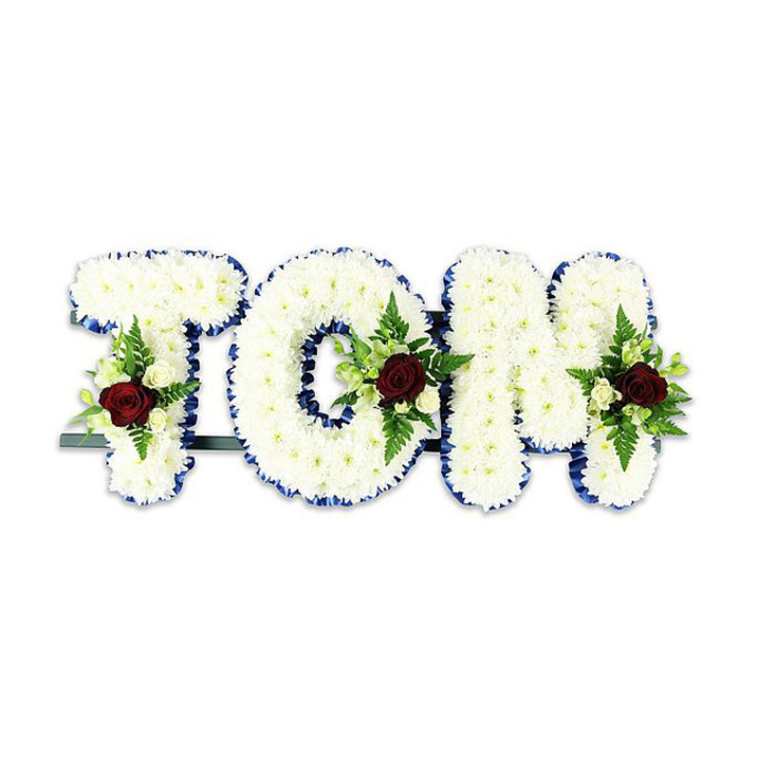 A personalized sympathy tribute spelling TOM, presented as large, three-dimensional letters densely covered in tiny, snow-white chrysanthemum-like flowers that form a soft, textured surface. The chrysanthemums are arranged so closely that the letters appear plush and substantial, each petal catching the light to create a gentle sheen across the forms. A vivid royal blue ribbon traces the exact edge of each letter, the crisp blue line providing a formal contrast that sharpens the silhouette and conveys a composed, respectful tone. Centered within each letter is a restrained floral spray: a single, deep red rose takes centre stage in every cluster, symbolising enduring love and remembrance, while small ivory spray roses and slim fern fronds weave around it to provide delicate highlights and fine, green texture. The combination of velvety red, creamy white and fresh green produces a balanced, classical palette that reads as both mournful and affectionate. Beneath the letters a thin dark metal support is visible, discreet but necessary for upright display at a chapel or graveside. The arrangement sits against a plain white background under soft, even light that emphasizes the tonal contrasts and the ribbons' crisp edges. Designed with the quiet knowledge of local needs in mind, this tribute would be equally fitting for memorials in Surrey Quays, near the shopping centre or along the Thames-facing docks, offering a handcrafted, measured expression of sympathy and honour.