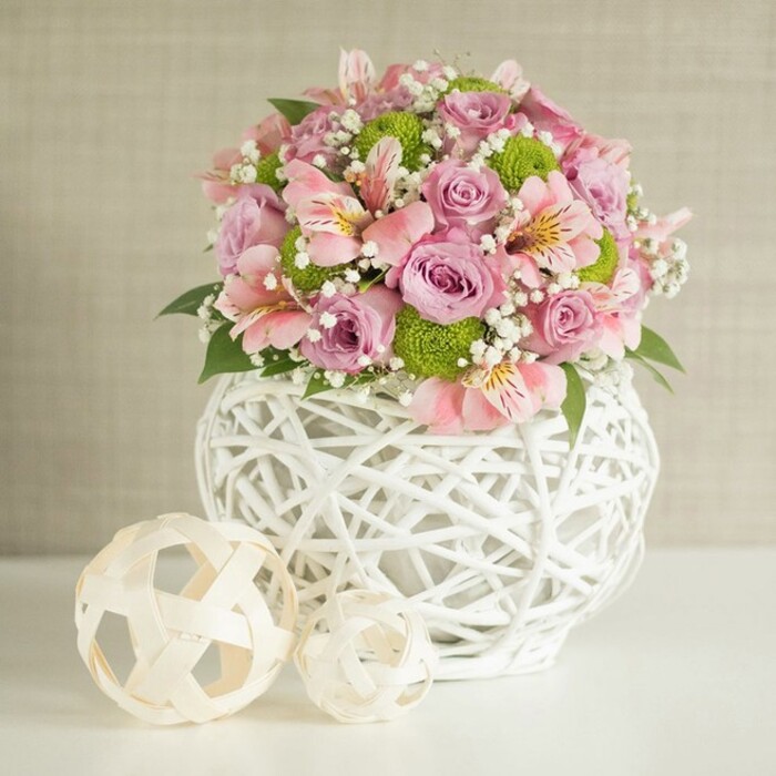 In this thoughtfully crafted floral centerpiece, the artistic flair of a local Surrey Quays florist shines through. Centred in the image, a sumptuous blend of soft pinks, lavenders, and electric lime green bursts elegantly from a unique, white statement vase reminiscent of the riverside district's stylish modern decor. The vase's openwork lattice, fashioned from intertwining white strips, evokes the airy feel of nearby waterside promenades and new developments, offering a contemporary twist on traditional craftsmanship. The bouquet itself brims with full, velvety pink and lavender roses, their delicate petals unfurling with quiet confidence around salmon-toned alstroemeria, which display slender, speckled petals and a hint of golden yellow at their core. Sprays of vivid lime green button chrysanthemums punctuate the arrangement with texture and lively visual rhythm; all around, the snowy delicacy of baby's breath adds a sense of gentle movement, like morning mist wafting over the Thames. Sprigs of dark green foliage add grounding depth, drawing every eye to the blooms' lushness. Completing the scene, two decorative spheres-woven from light, natural fiber-rest nearby, their organic forms echoing both the vase's artistry and the community's love of natural detail. Bathed in soft, ambient light against a subtly textured beige background, this composition feels perfect for elegant weddings, at-home celebrations, or simply brightening a serene Surrey Quays afternoon.