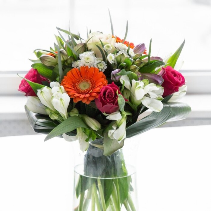 This meticulously crafted Surrey Quays bouquet is a testament to the harmonious interplay of colour, texture, and local artistry. At its core, creamy and peach gerberas fan outward, their petals soft and warm-radiating like the gentle sunrise over South Dock Marina. Fiery orange gerberas punctuate the arrangement, infusing energy reminiscent of the vibrant spirit that animates Lower Road. Nestled among them, luxurious fuchsia roses spiral outwards, exuding depth and an understated opulence. White alstroemeria and finely clustered spray roses introduce luminous highlights, softening the effect and evoking the tranquillity of green walking paths nearby. Broad, glossy aspidistra leaves arch protectively around the bouquet's base, grounding the kaleidoscope of blooms with their bold verdant curves. Smaller pointed leaves and feathery greenery fill the spaces, creating subtle layers suggestive of a lush riverside garden. Light, possibly from a sunlit window, bathes the arrangement, enhancing the colours' vibrancy while honoring their natural character. As the bouquet sits in a clear vase, the stems stand tall and visible-a final touch of freshness and authenticity. Whether marking a joyful gathering, a new home near Surrey Quays station, or a thoughtful thank you, this arrangement brings optimism and sophistication, echoing the creative flair of Flowers Surrey Quays.