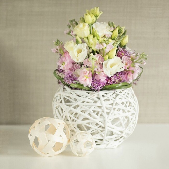 A stunningly curated floral arrangement, distinctly shaped by the expert touch of a Surrey Quays florist, radiates natural grace and gentle luxury. Overflowing with plush eustoma in creamy white and pastel yellow, the bouquet is further enriched by towers of soft pink matthiola, their blossoms arranged with artistic precision for maximum depth and movement. Textural intrigue comes from the dense clusters of deep purple filler-evocative of asters or small allium-carefully nestled at the bouquet's base, while a fresh band of dark green foliage provides a seamless circular frame. All are cradled in an open-weave, spherical white vase, handcrafted from intertwined rattan or painted twigs, revealing glimpses of the inner mechanics and creating a striking interplay of light and shadow. Positioned atop a luminous white tabletop, the arrangement is complemented by two sculptural accent spheres made of light wood or woven reed, echoing the vase's artisanal quality and referencing Surrey Quays' tradition of craft markets. The softly illuminated setting is finished by a subtle vertical fabric-textured wall in warm beige, which recedes quietly to allow the bouquet's pastel tones to shine. This image not only conveys themes of renewal, elegance, and celebration, but also captures the spirit of Surrey Quays living-where waterfront calm, creative flair, and fresh, seasonal flowers converge for weddings, gatherings, or moments of tranquil beauty at home.