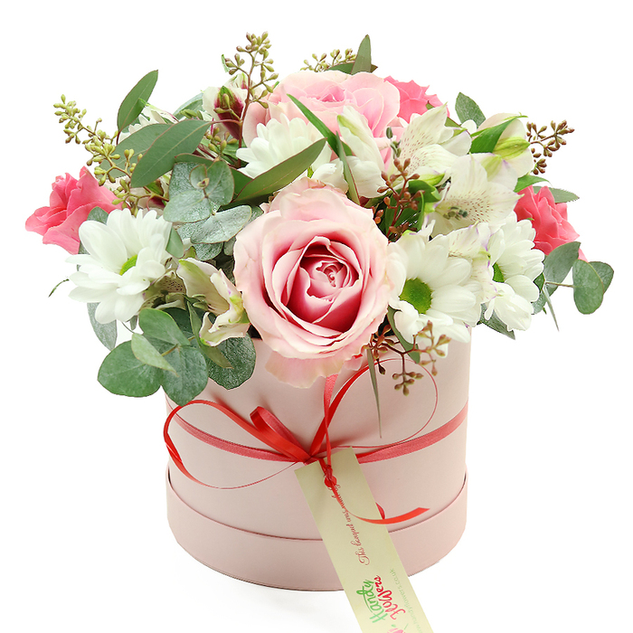Set against a pristine white backdrop, a chic pastel pink hatbox brimming with flowers becomes the clear centre of attention, its simple cylindrical shape and matte finish lending a refined, contemporary feel. A slim coral-red satin ribbon circles the box and is tied into a tidy bow at the front, where a cream-coloured gift tag with soft green script gently dangles, implying a personal message or dedication ready to be delivered to a loved one in Surrey Quays. From this understated base, the bouquet rises in a generous dome, overflowing in soft pinks, whites, and greens. Several light pink roses, some with deeper fuchsia swirls at their cores, anchor the design; one especially large bloom faces outward, fully open and layered with velvety petals that suggest both tenderness and quiet luxury. Around and between these roses sit white daisy-like chrysanthemums, each flower a perfect circle of crisp white petals encasing a yellow-green centre that adds tiny flashes of brightness, like sunlight catching the water at nearby South Dock. White alstroemeria, easily recognised by their slender, trumpet-shaped petals and delicate speckles, thread through the arrangement, while smaller, richer pink blossoms-perhaps spray roses or lisianthus-introduce pockets of intense colour to enliven the pastel palette. A thoughtful mix of greenery frames the flowers: silvery eucalyptus with rounded leaves, darker green foliage, and fine sprigs tipped with buds and tiny brownish seed heads, giving the composition depth and texture. The lighting is even and soft, enhancing every petal edge without distraction. The result is a polished, romantic hatbox arrangement that feels perfect for an anniversary dinner in a Surrey Quays apartment, a Mother's Day surprise, or a heartfelt thank-you delivered locally with care.