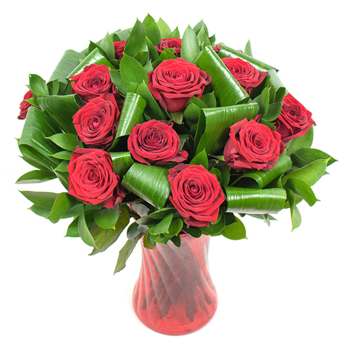A luxurious, expertly balanced bouquet of a dozen plus deep crimson roses is captured here, every bloom opened to reveal the velvety, spiral layers of petals that give each head a plush, tactile quality. The roses form a compact, rounded crown, their intense red tones amplified by soft, even lighting and set starkly against a pristine white background so that colour and texture become the sole focus. Rich, dark green foliage is used abundantly as both backdrop and structural element: smaller, glossy leaves - reminiscent of salal or ruscus - provide a dense, verdant cushion, while larger, lustrous leaves have been curled and folded into sculptural tubes and arcs that cradle and frame the roses with an architectural grace. This interplay of soft floral surfaces and glossy, folded greenery gives the arrangement a contemporary silhouette while retaining classic romantic symbolism. The bouquet rests in a tall, translucent ruby-red vase with a subtle twisted-rib texture; its slender waist and flared mouth mirror the bouquet's proportions and reflect vertical light lines that heighten the composition's elegance. Imagine the faint, nostalgic perfume of freshly opened roses lifting from the arrangement, a scent that evokes anniversary dinners or refined Valentine's moments. As a florist serving Surrey Quays and nearby Rotherhithe, I see this arrangement working beautifully as a centerpiece for intimate celebrations or as a considered same-day delivery for heartfelt gestures around Greenland Dock and Canada Water.