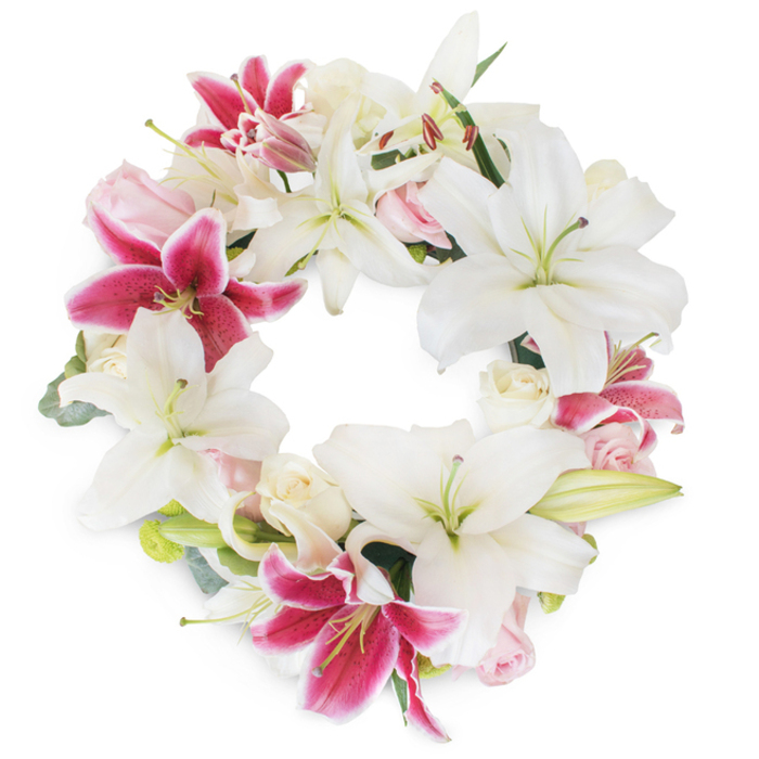 A radiant and meaningful floral wreath, designed with thoughtful sensitivity for memorial occasions in Surrey Quays, stands out against a spotless white background. At its heart, an abundant array of white oriental lilies fans out into wide, elegant trumpets, their broad petals subtly curled and highlighted by soft green pistils and creamy stamens. These radiant whites invoke a sense of tranquility and purity, echoing the peaceful mornings spent along the quaysides and communal gardens of Surrey Quays. Accentuating the elegant display are dramatic fuchsia-pink lilies-each bloom infused with vibrant magenta and delicate inner markings-creating bold focal points and a striking harmony within the composition. Amid the lilies, delicate blush-pink and ivory roses-some unfurling in full, others mere buds-soften the design with their romantic texture, mirroring the quiet beauty often found in local residential gardens. Small hints of eucalyptus and muted greenery are skillfully threaded through the circumference, lending structural balance and a touch of woodland freshness evocative of nearby Russia Dock Woodland. The precise, circular arrangement of the wreath, with its open center and generous layering of florals, embodies completeness, unity, and remembrance. This floral creation, carefully arranged for the Surrey Quays community, is not only visually captivating but emits an air of sincere compassion, making it suited for both private moments of reflection and dignified celebrations of life.