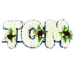 A personalised floral tribute spells out the name TOM in large, freestanding letters, crafted with the care and precision you might expect from an experienced Surrey Quays funeral florist. Each letter is formed from a bed of snow-white chrysanthemums, packed tightly together so that their rounded heads create a plush, uniform surface. The white blooms catch the light softly, giving the arrangement a gentle luminosity against the plain white background. Around the perimeter of every letter, a royal blue satin ribbon has been artfully pleated, providing a crisp, elegant outline that enhances the clarity of the name and adds a sense of quiet dignity. At select points on the T, O, and M, clusters of accent flowers draw the eye: deep, velvety red roses are the main focus, their rich colour symbolising love and remembrance. They are surrounded by smaller white or cream spray roses and slender white freesia-like blooms, all nestled into a backdrop of lush green foliage and lacy fern. These touches of red and green break up the expanse of white chrysanthemums, introducing depth, contrast, and a natural, garden-like feel. The letters are mounted on a narrow dark frame so they align neatly in a row, imagined perhaps resting at a crematorium chapel near Canada Water or on a graveside in a quiet Surrey Quays cemetery. The overall impression is one of composed elegance and sincerity, a traditional yet deeply personal floral arrangement created to honour Tom's memory and offer comfort to those gathered to mourn.