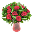 A sculpted bouquet of a dozen deep crimson roses sits poised in a tall ruby-red glass vase, its velvety petals unfurling in full, perfect blooms that read like a quiet declaration of love. The roses are arranged into a lush, rounded head, each flower's spiral of tightly wound petals opening to reveal the satiny, wine-dark interiors that catch the light and seem to glow from within. Surrounding and cradling each bloom is a generous halo of glossy, dark green foliage - broad salal- or ruscus-like leaves form a dense backdrop while larger, emerald-hued leaves are meticulously rolled and curved into elegant loops and tubes, adding sculptural arches that bring architectural poise to the composition. Soft, even lighting removes harsh shadows and sets the reds and greens against a crisp white studio background so every texture - the tender velvet of the roses, the sheen of the greens, and the subtle ribbed twist of the glass - is clearly visible. The vase's translucent ruby tone and vertical swirl pattern echo the roses' passionate hue and introduce slender, reflective lines that lead the eye upward, giving the arrangement an elongated, refined silhouette. Close your eyes and imagine the classic, faint perfume of red roses mingling with the cool green scent of fresh foliage - calm, romantic, and timeless. This bouquet reads as a luxury gift or centerpiece for anniversaries, Valentine's moments, or an intimate dinner, a floral language of deep devotion crafted with considered structure and modern flair, well suited to celebrations across Surrey Quays and nearby Canada Water.