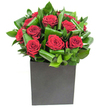 In this elegantly modern composition, a cluster of deep crimson roses takes centre stage, each bloom opening in velvety spirals that catch the light with a soft, plush sheen. The roses are tightly arranged and tucked into broad, glossy green leaves that have been artfully curled and looped around the stems, creating a steady rhythm of form and texture that feels both sculptural and organic. The foliage's waxy surfaces throw back highlights under a clear, studio-like light, giving the greenery a lively, almost tropical freshness that contrasts with the warm, dense red of the petals. The bouquet sits in a matte black, square cube-its hard-edged, understated base grounding the composition and amplifying the colours above, the flat finish quietly absorbing light so the blooms appear to float. A subtle fragrance seems to rise from the arrangement: the classic, heady sweetness of rose, brightened by cool, leafy notes like a breath of fresh air from a nearby dock. I imagine placing this piece on a contemporary dining table in a Surrey Quays apartment or as a thoughtful centrepiece beside Greenland Dock, where the crispness of river air would echo the bouquet's clean lines. The overall effect is poised and confident-a modern declaration of romance or sincere gratitude crafted with exacting care by a local hand. Every curl of leaf and every spiral of petal reads as an intentional choice, designed to make an intimate space feel composed, celebratory, and quietly memorable.